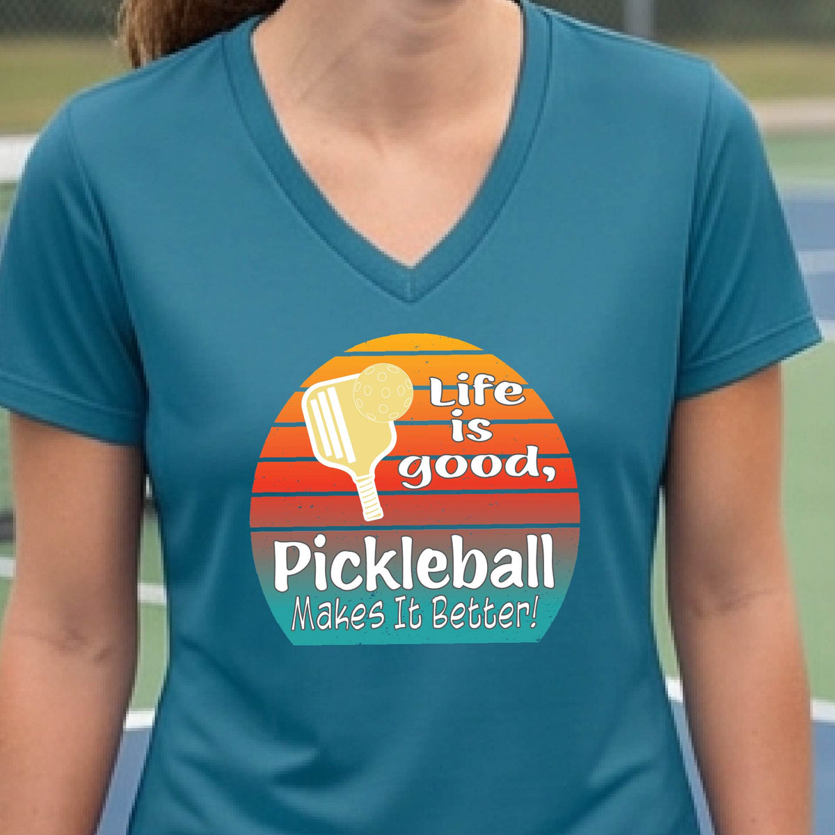 A person wearing a teal blue V-neck t-shirt with a retro-style pickleball graphic on the chest. The graphic features a yellow pickleball paddle, a red, orange, and yellow sunset gradient, and the text 'Life is good, Pickleball Makes It Better!'.