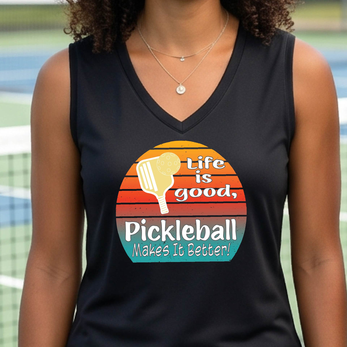 A  woman wearing a black short-sleeve t-shirt with a retro multi-colored circle with a pickleball paddle and ball, with white text that says 'LIFE IS GOOD PICKLEBALL MAKES IT BETTER', standing in front of a pickleball court.