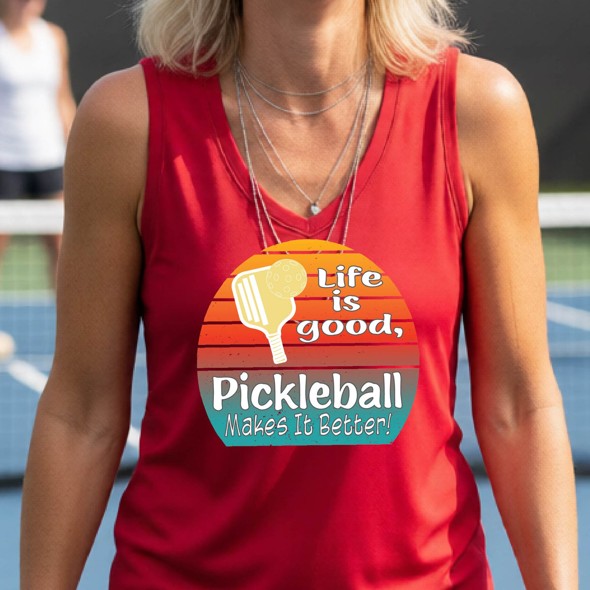 A  woman wearing a red short-sleeve t-shirt with a retro multi-colored circle with a pickleball paddle and ball, with white text that says 'LIFE IS GOOD PICKLEBALL MAKES IT BETTER', standing in front of a pickleball court.