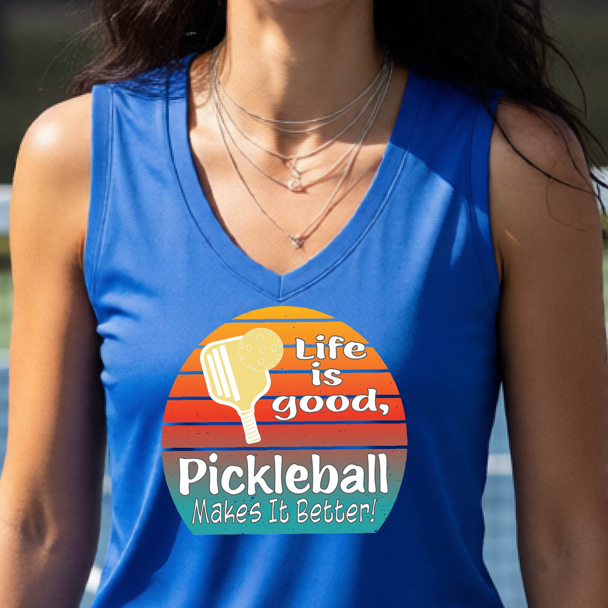 A  woman wearing a royal blue short-sleeve t-shirt with a retro multi-colored circle with a pickleball paddle and ball, with white text that says 'LIFE IS GOOD PICKLEBALL MAKES IT BETTER', standing in front of a pickleball court.