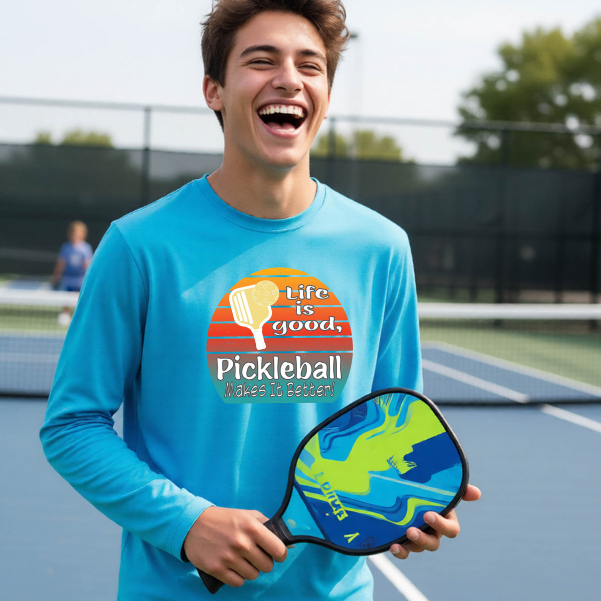 A youth wearing a bright blue short-sleeve t-shirt with a retro multi-colored circle with a pickleball paddle and ball, with white text that says 'LIFE IS GOOD PICKLEBALL MAKES IT BETTER', standing in front of a pickleball court.