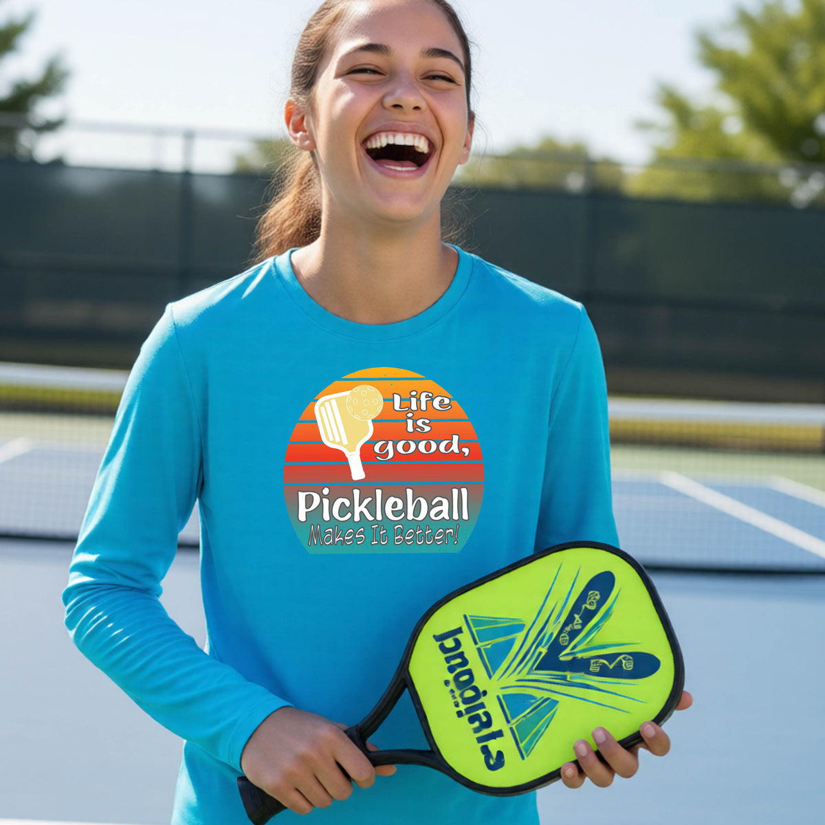 A youth wearing a bright blue short-sleeve t-shirt with a retro multi-colored circle with a pickleball paddle and ball, with white text that says 'LIFE IS GOOD PICKLEBALL MAKES IT BETTER', standing in front of a pickleball court.