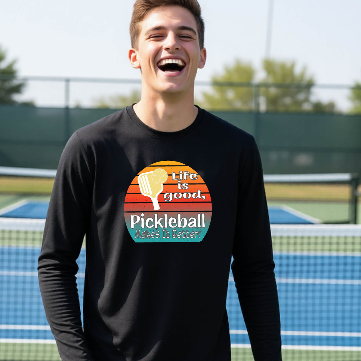 A youth wearing a black short-sleeve t-shirt with a retro multi-colored circle with a pickleball paddle and ball, with white text that says 'LIFE IS GOOD PICKLEBALL MAKES IT BETTER', standing in front of a pickleball court.