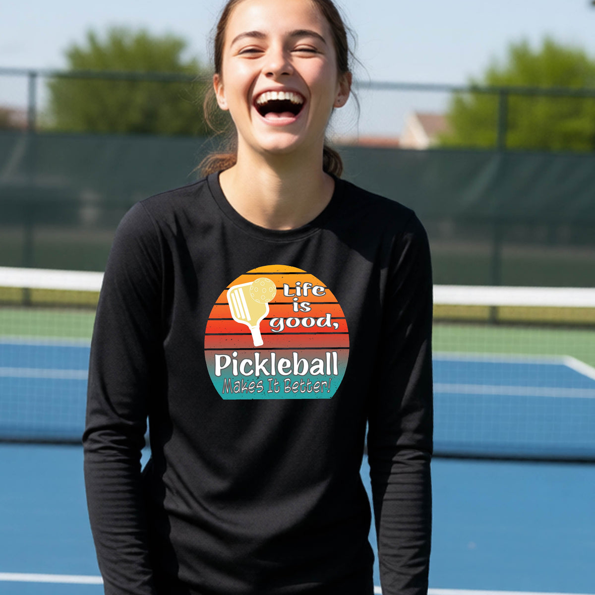 A youth wearing a black short-sleeve t-shirt with a retro multi-colored circle with a pickleball paddle and ball, with white text that says 'LIFE IS GOOD PICKLEBALL MAKES IT BETTER', standing in front of a pickleball court.