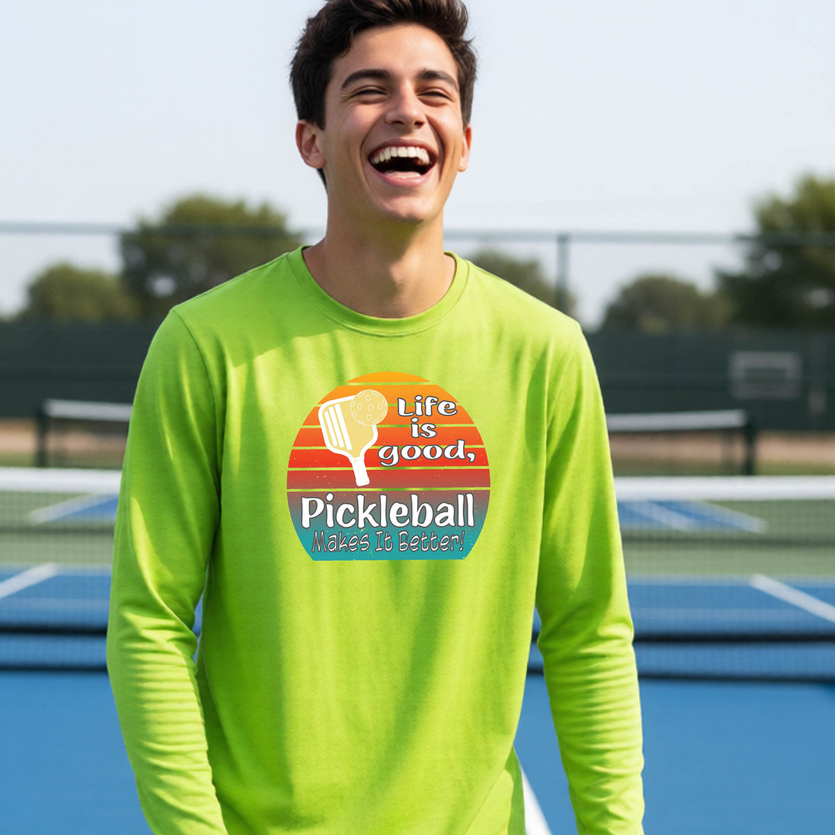 A youth wearing a lime green short-sleeve t-shirt with a retro multi-colored circle with a pickleball paddle and ball, with white text that says 'LIFE IS GOOD PICKLEBALL MAKES IT BETTER', standing in front of a pickleball court.