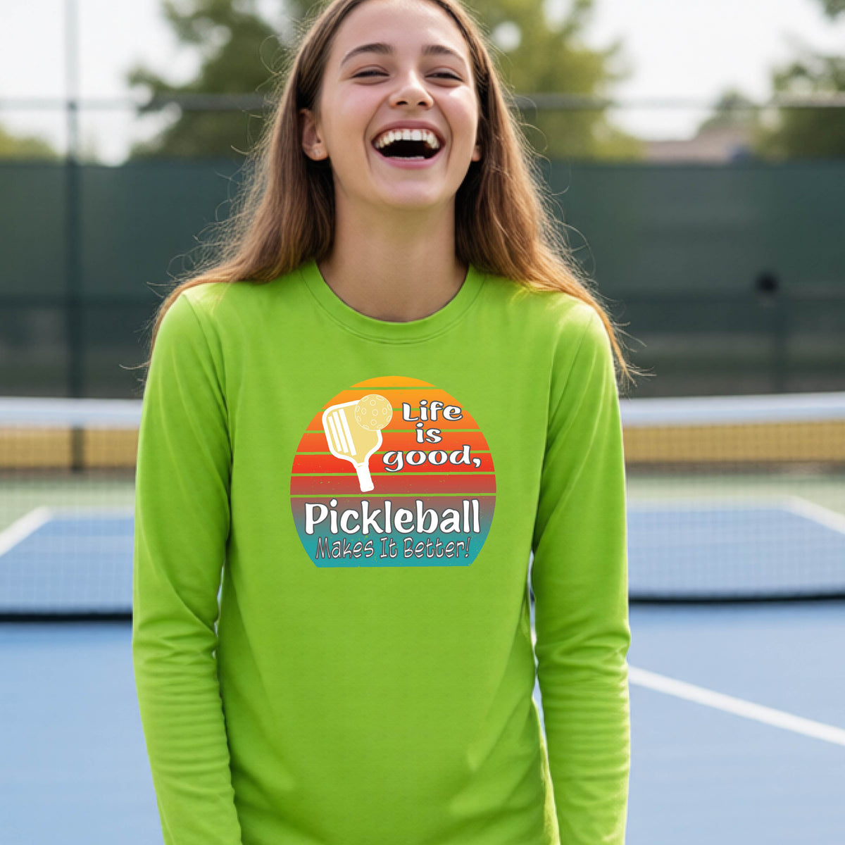 A youth wearing a lime green short-sleeve t-shirt with a retro multi-colored circle with a pickleball paddle and ball, with white text that says 'LIFE IS GOOD PICKLEBALL MAKES IT BETTER', standing in front of a pickleball court.