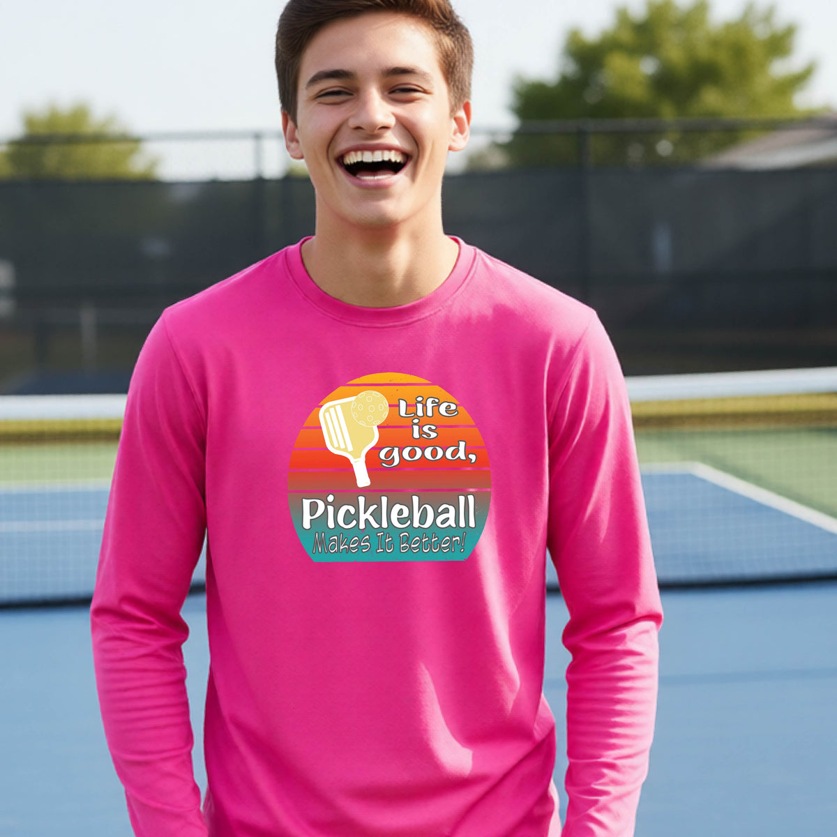 A youth wearing a pink short-sleeve t-shirt with a retro multi-colored circle with a pickleball paddle and ball, with white text that says 'LIFE IS GOOD PICKLEBALL MAKES IT BETTER', standing in front of a pickleball court.