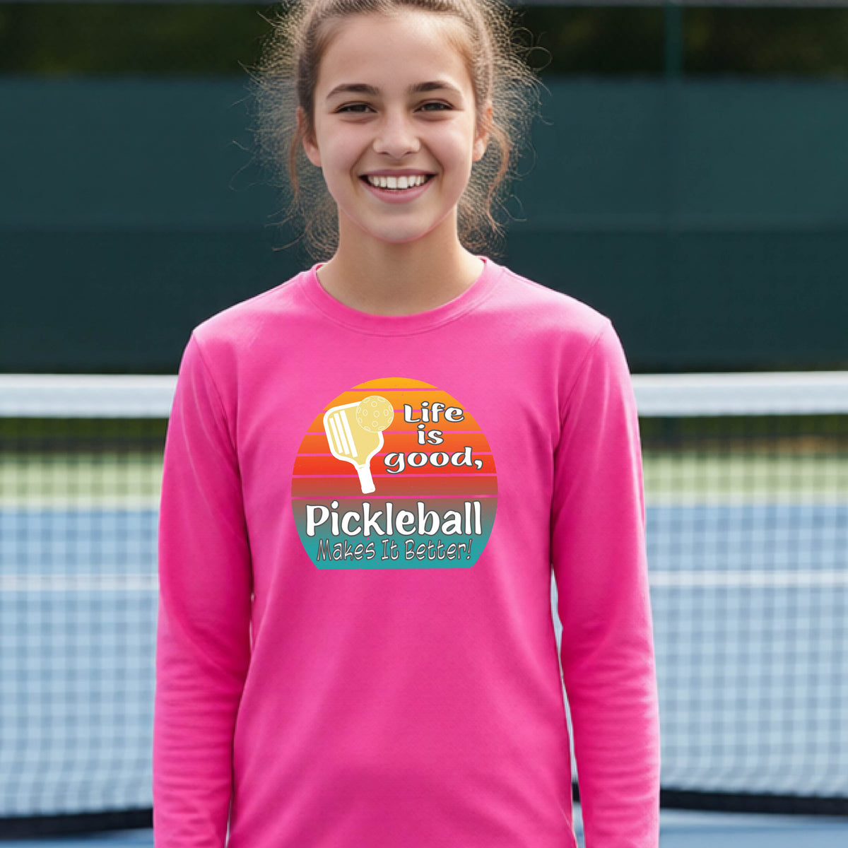 A youth wearing a pink short-sleeve t-shirt with a retro multi-colored circle with a pickleball paddle and ball, with white text that says 'LIFE IS GOOD PICKLEBALL MAKES IT BETTER', standing in front of a pickleball court.