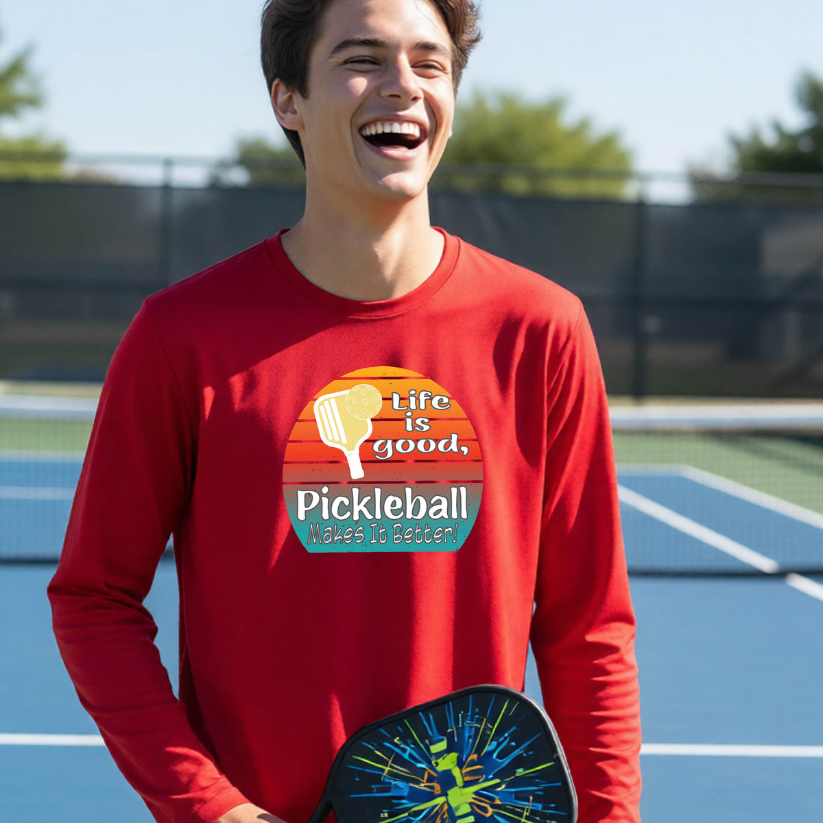 A youth wearing a red short-sleeve t-shirt with a retro multi-colored circle with a pickleball paddle and ball, with white text that says 'LIFE IS GOOD PICKLEBALL MAKES IT BETTER', standing in front of a pickleball court.