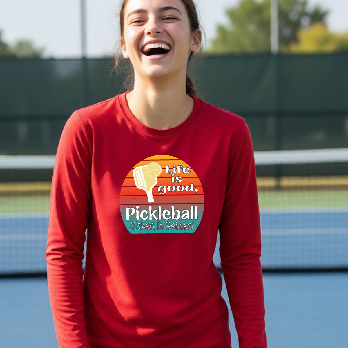 A youth wearing a red short-sleeve t-shirt with a retro multi-colored circle with a pickleball paddle and ball, with white text that says 'LIFE IS GOOD PICKLEBALL MAKES IT BETTER', standing in front of a pickleball court.