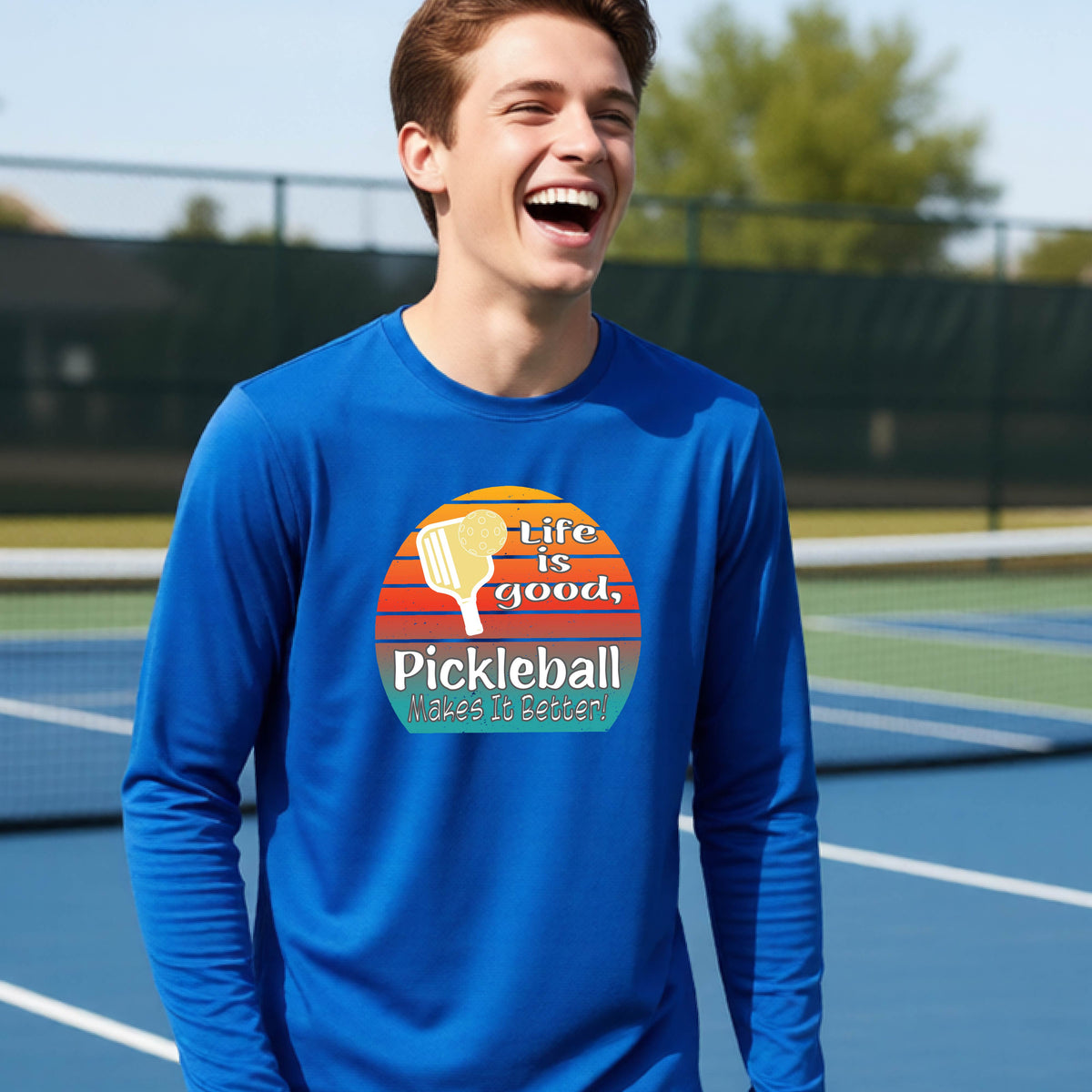 A youth wearing a royal blue short-sleeve t-shirt with a retro multi-colored circle with a pickleball paddle and ball, with white text that says 'LIFE IS GOOD PICKLEBALL MAKES IT BETTER', standing in front of a pickleball court.