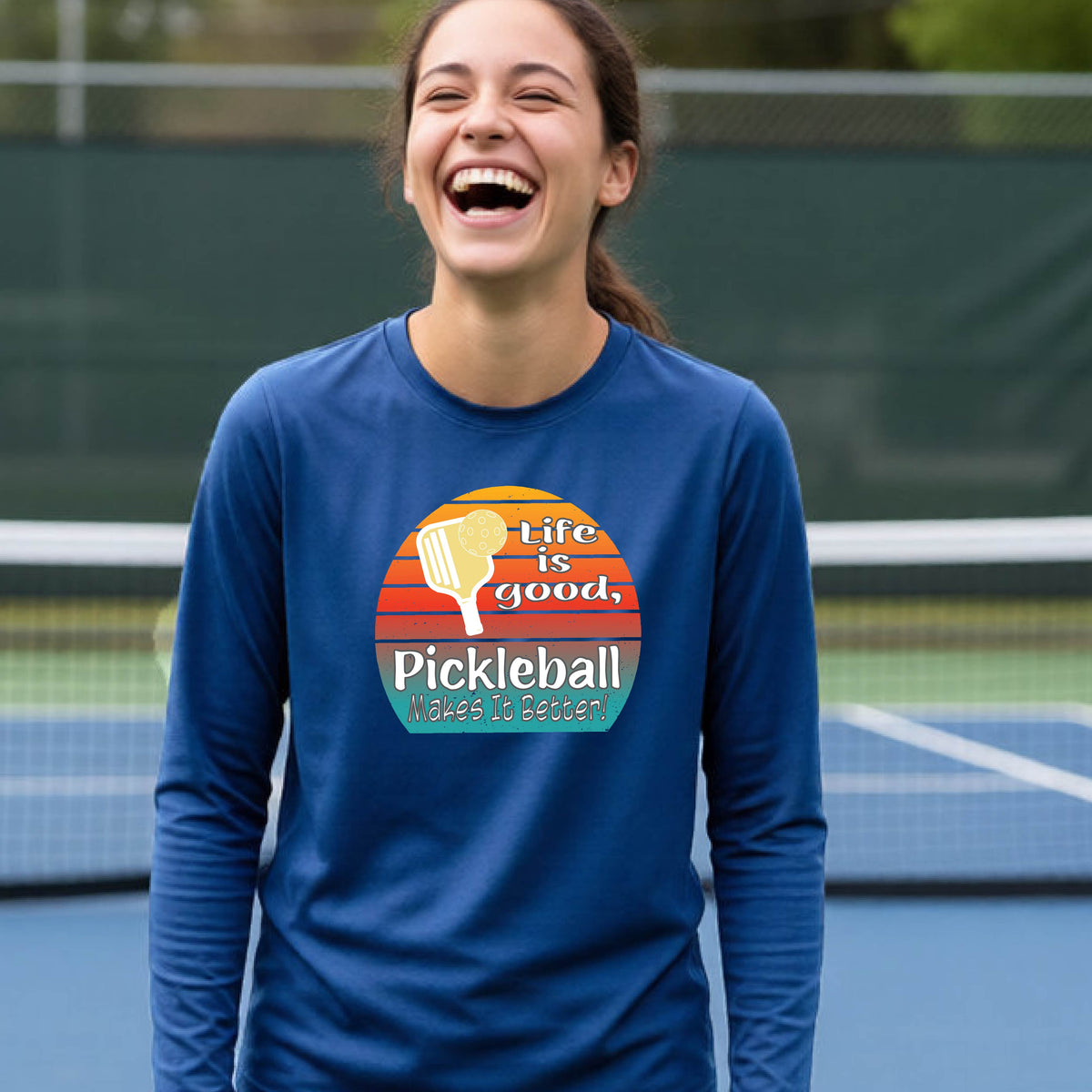 A youth wearing a royal blue short-sleeve t-shirt with a retro multi-colored circle with a pickleball paddle and ball, with white text that says 'LIFE IS GOOD PICKLEBALL MAKES IT BETTER', standing in front of a pickleball court.