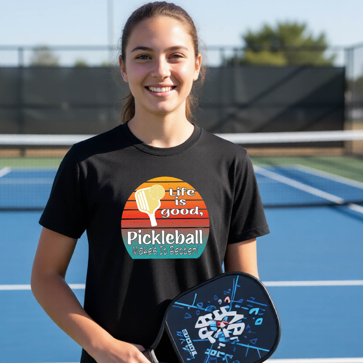 A teenager wearing a black short-sleeve t-shirt with a retro multi-colored circle with a pickleball paddle and ball, with white text that says 'LIFE IS GOOD PICKLEBALL MAKES IT BETTER', standing in front of a pickleball court.