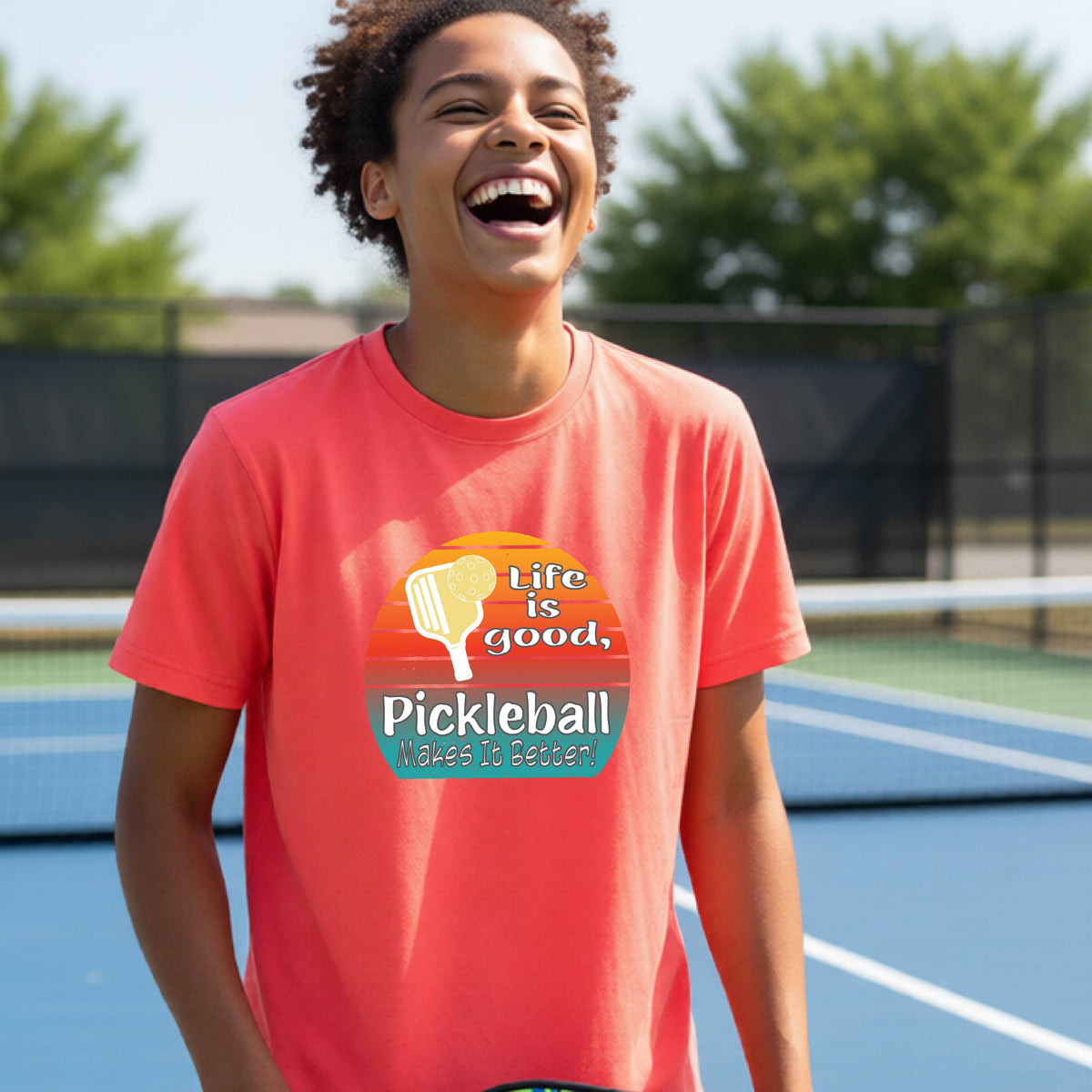 A teenager wearing a coral pink short-sleeve t-shirt with a retro multi-colored circle with a pickleball paddle and ball, with white text that says 'LIFE IS GOOD PICKLEBALL MAKES IT BETTER', standing in front of a pickleball court.