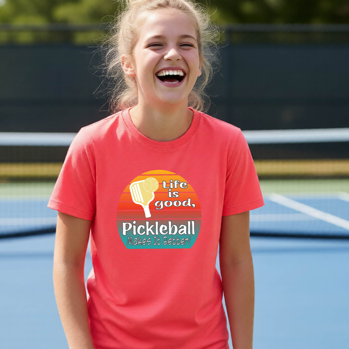 A teenager wearing a coral pink short-sleeve t-shirt with a retro multi-colored circle with a pickleball paddle and ball, with white text that says 'LIFE IS GOOD PICKLEBALL MAKES IT BETTER', standing in front of a pickleball court.