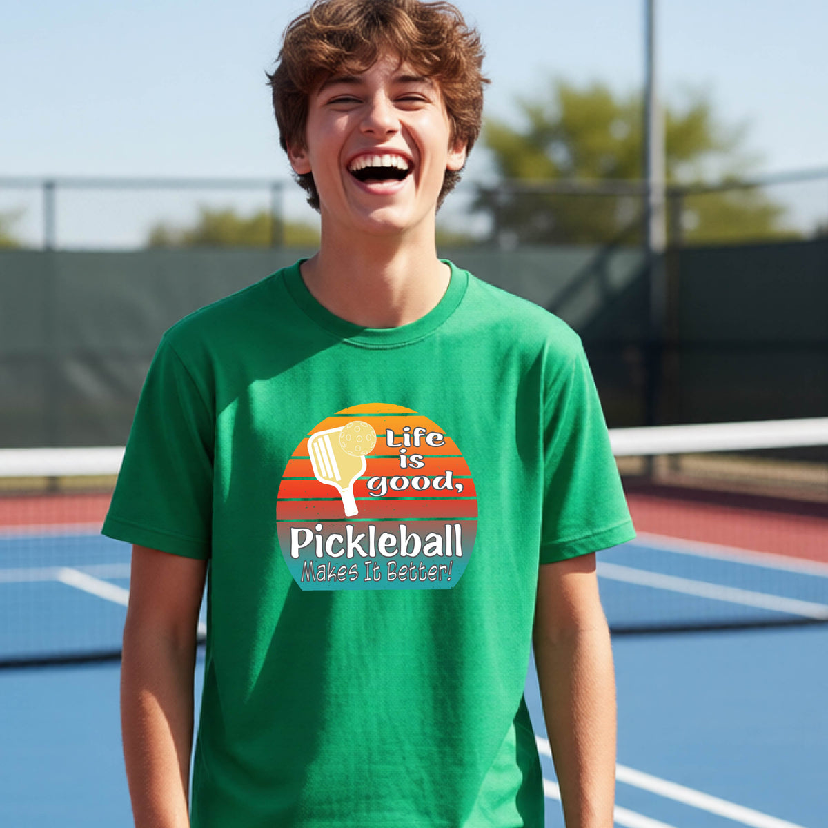 A teenager wearing a green short-sleeve t-shirt with a retro multi-colored circle with a pickleball paddle and ball, with white text that says 'LIFE IS GOOD PICKLEBALL MAKES IT BETTER', standing in front of a pickleball court.