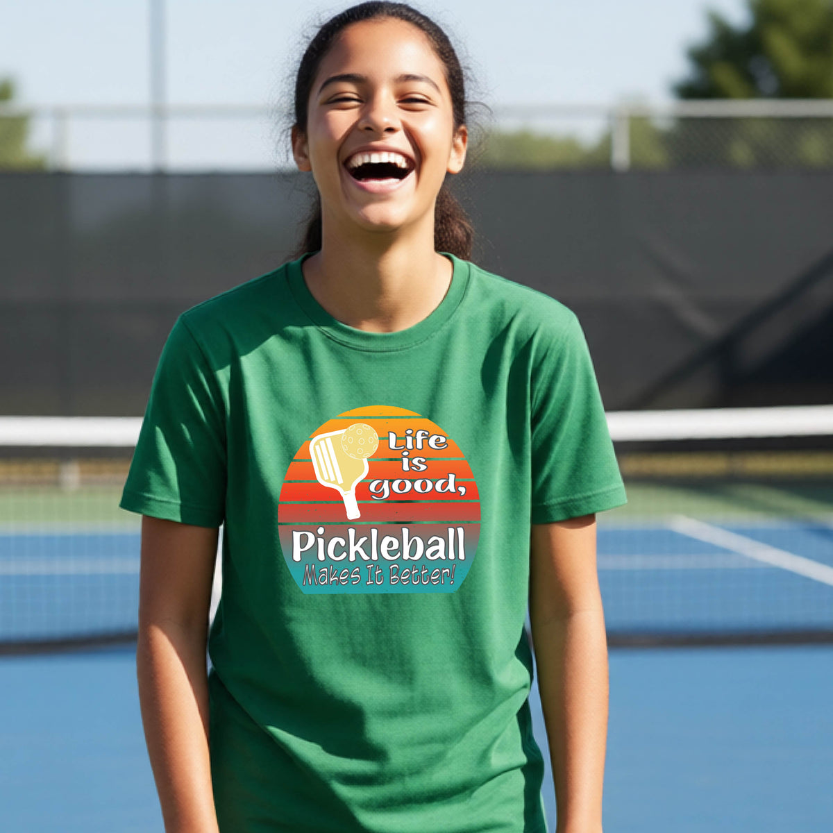 A teenager wearing a green short-sleeve t-shirt with a retro multi-colored circle with a pickleball paddle and ball, with white text that says 'LIFE IS GOOD PICKLEBALL MAKES IT BETTER', standing in front of a pickleball court.
