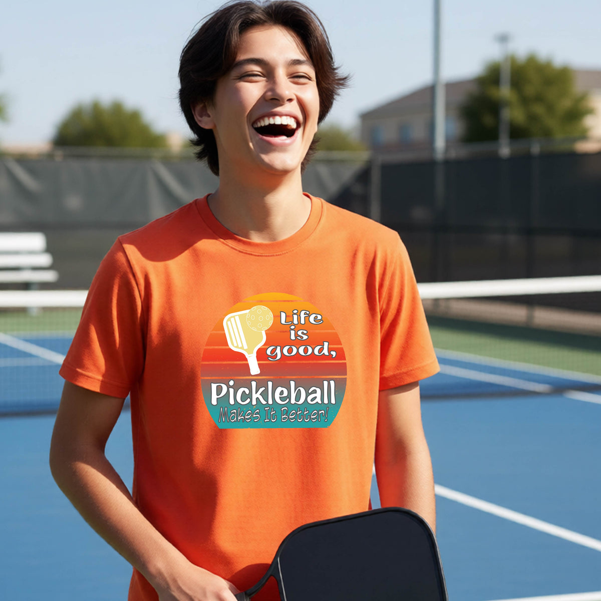A teenager wearing an orange short-sleeve t-shirt with a retro multi-colored circle with a pickleball paddle and ball, with white text that says 'LIFE IS GOOD PICKLEBALL MAKES IT BETTER', standing in front of a pickleball court.