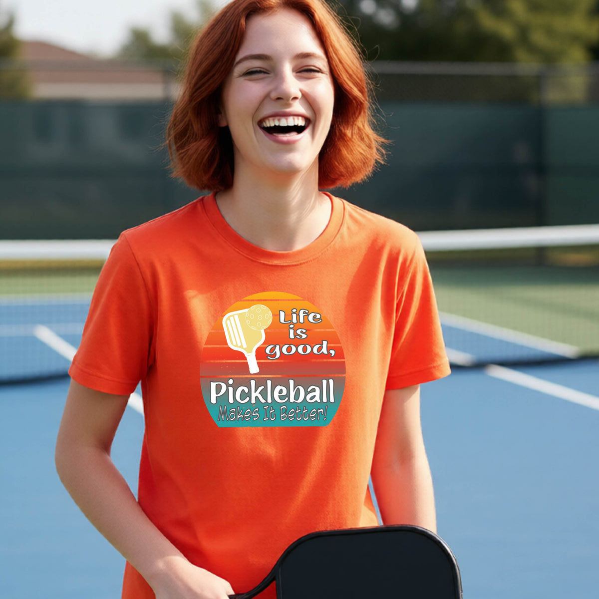 A teenager wearing an orange short-sleeve t-shirt with a retro multi-colored circle with a pickleball paddle and ball, with white text that says 'LIFE IS GOOD PICKLEBALL MAKES IT BETTER', standing in front of a pickleball court.