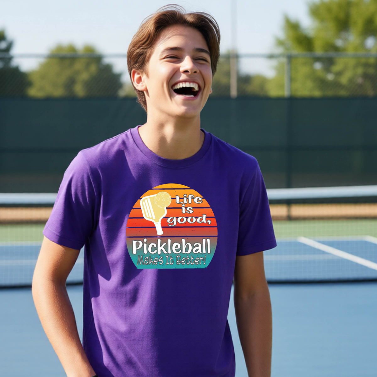 A teenager wearing a purple short-sleeve t-shirt with a retro multi-colored circle with a pickleball paddle and ball, with white text that says 'LIFE IS GOOD PICKLEBALL MAKES IT BETTER', standing in front of a pickleball court.