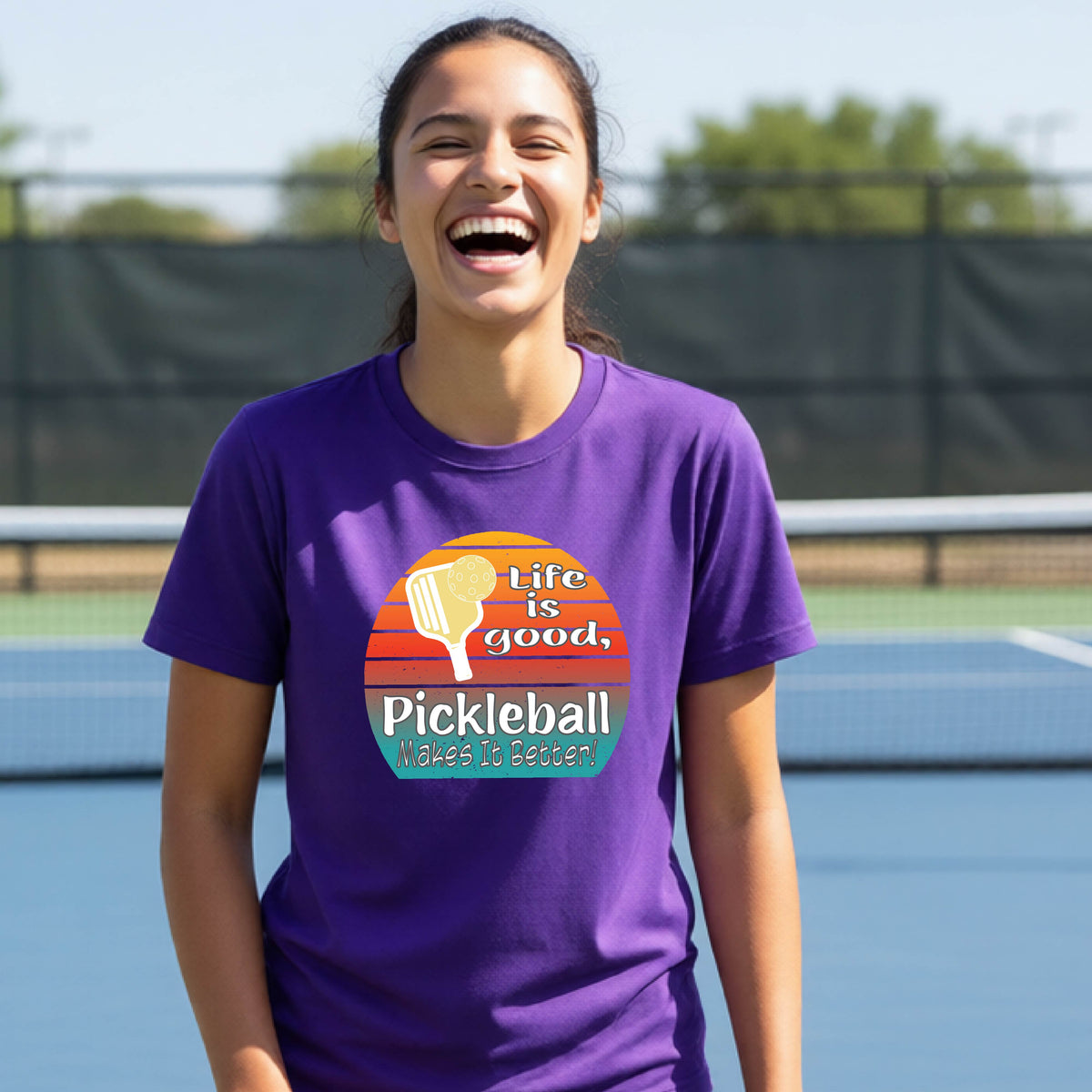 A teenager wearing a purple short-sleeve t-shirt with a retro multi-colored circle with a pickleball paddle and ball, with white text that says 'LIFE IS GOOD PICKLEBALL MAKES IT BETTER', standing in front of a pickleball court.