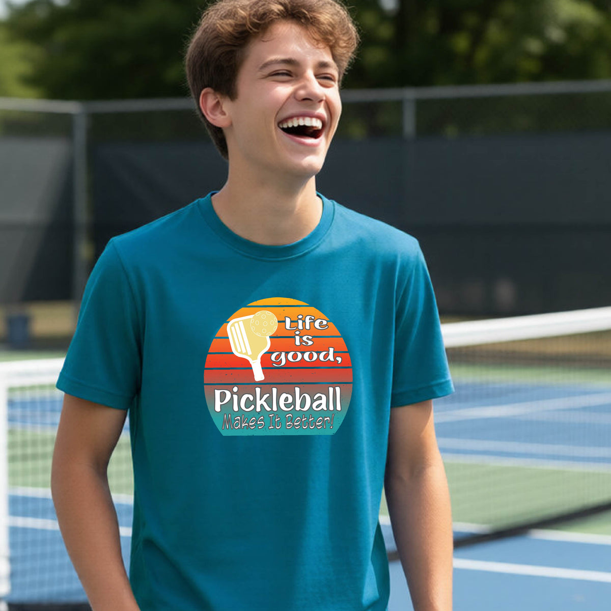 A teenager wearing a teal short-sleeve t-shirt with a retro multi-colored circle with a pickleball paddle and ball, with white text that says 'LIFE IS GOOD PICKLEBALL MAKES IT BETTER', standing in front of a pickleball court.