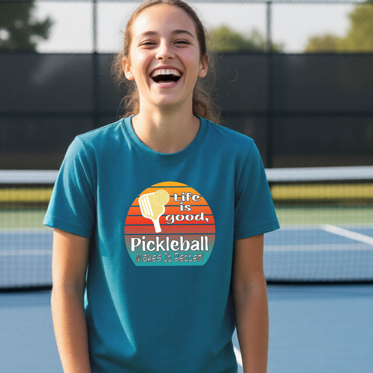 A teenager wearing a teal short-sleeve t-shirt with a retro multi-colored circle with a pickleball paddle and ball, with white text that says 'LIFE IS GOOD PICKLEBALL MAKES IT BETTER', standing in front of a pickleball court.