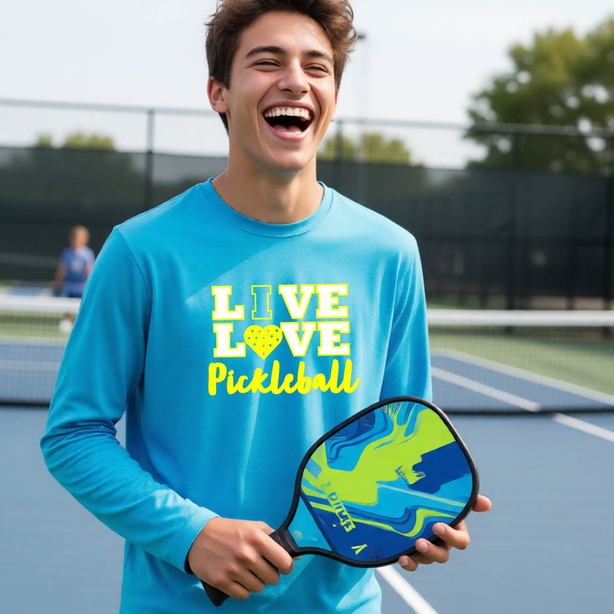 A teenager on a pickleball court wearing a bright blue long-sleeve V-neck shirt with a yellow graphic that reads 'LIVE LOVE Pickleball' and a heart-shaped pickleball dot pattern inside the word 'LOVE'.