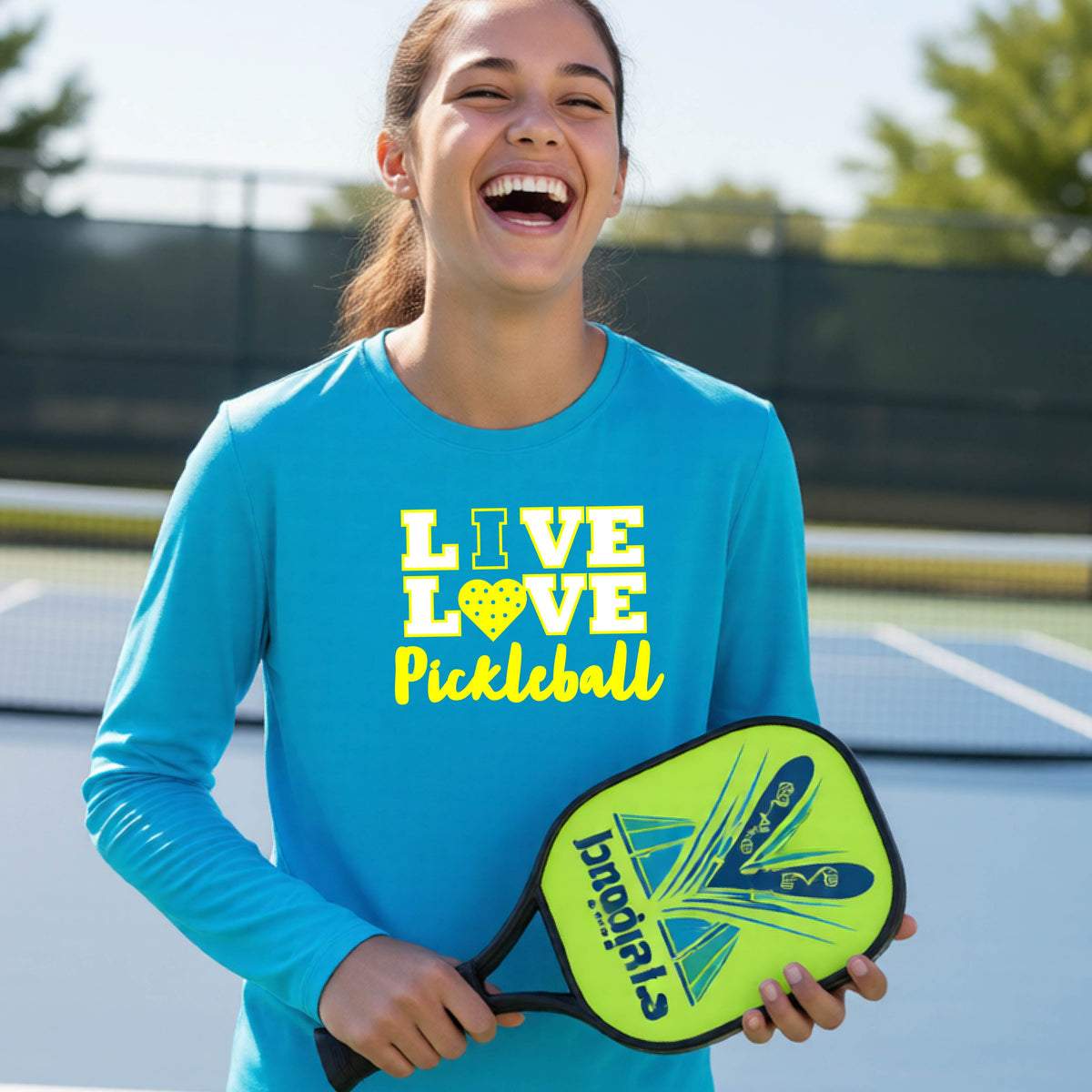 A teenager on a pickleball court wearing a bright blue long-sleeve V-neck shirt with a yellow graphic that reads 'LIVE LOVE Pickleball' and a heart-shaped pickleball dot pattern inside the word 'LOVE'.