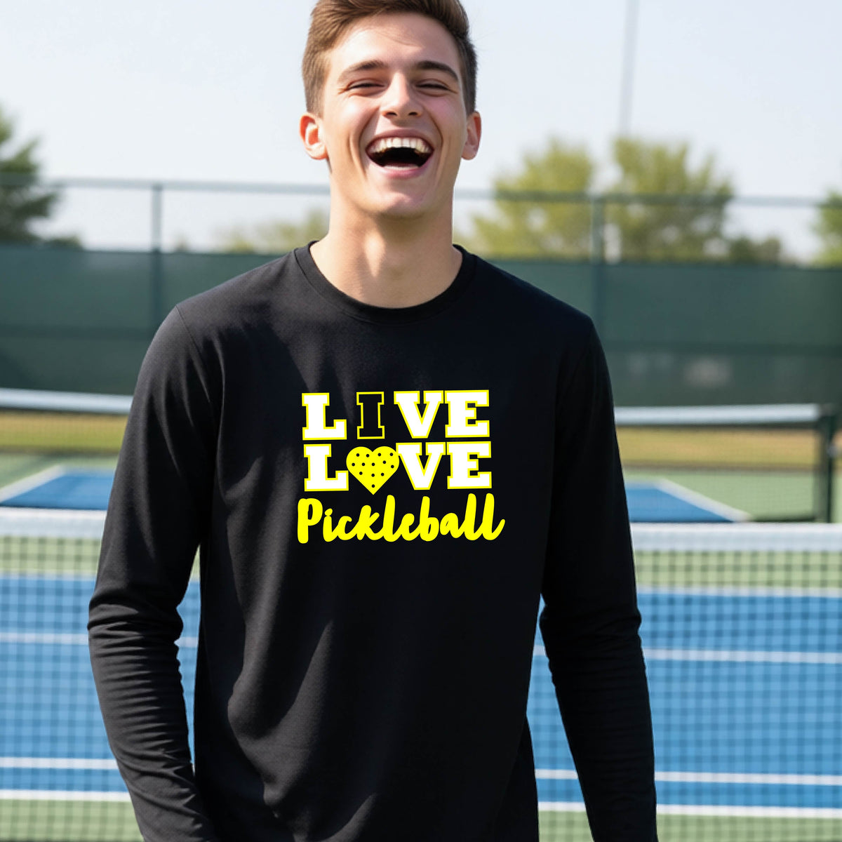 A teenager on a pickleball court wearing a black long-sleeve V-neck shirt with a yellow graphic that reads 'LIVE LOVE Pickleball' and a heart-shaped pickleball dot pattern inside the word 'LOVE'.