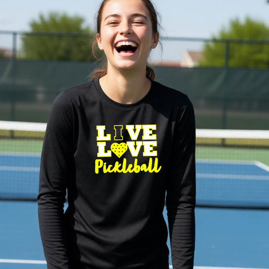 A teenager on a pickleball court wearing a black long-sleeve V-neck shirt with a yellow graphic that reads 'LIVE LOVE Pickleball' and a heart-shaped pickleball dot pattern inside the word 'LOVE'.