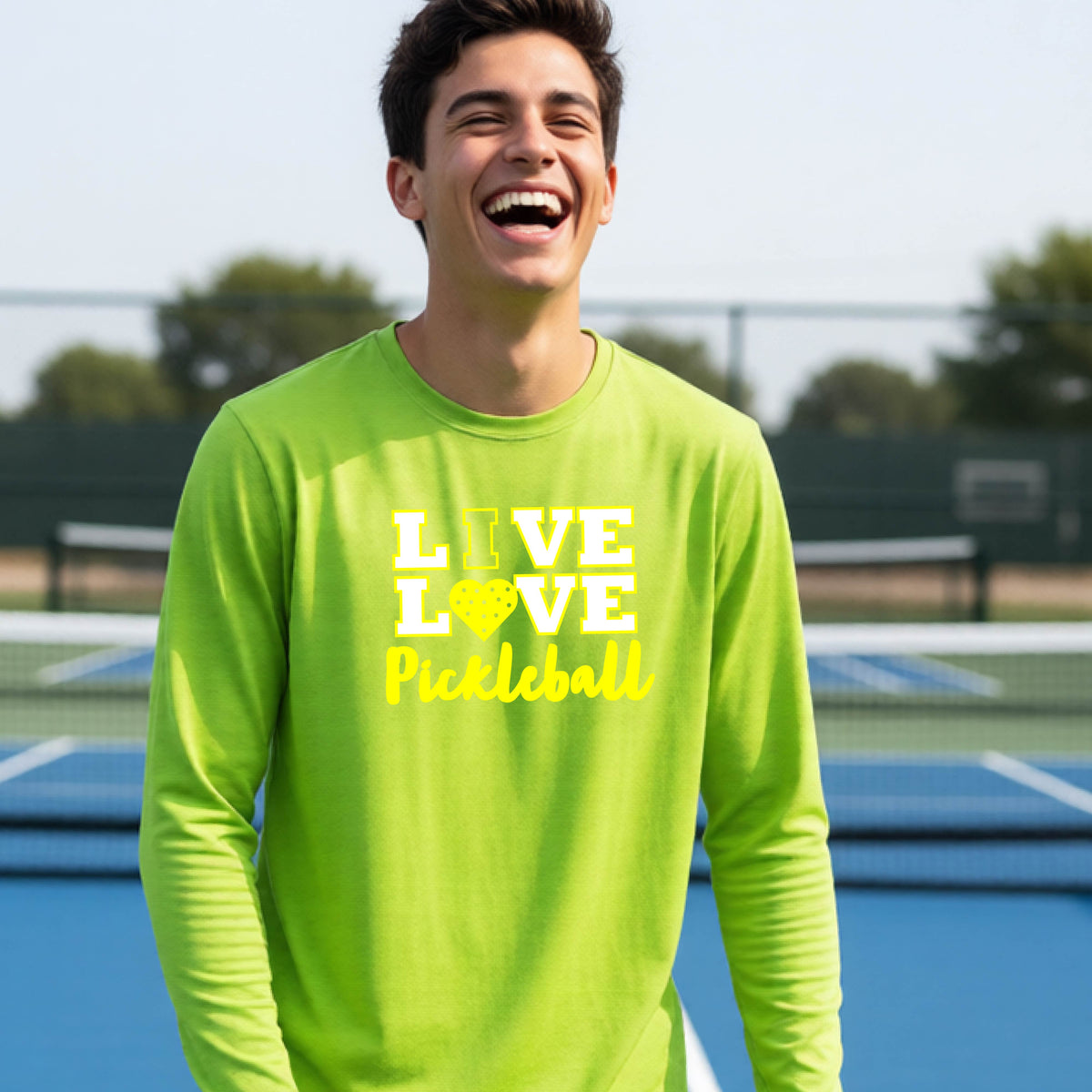 A teenager on a pickleball court wearing a lime green long-sleeve V-neck shirt with a yellow graphic that reads 'LIVE LOVE Pickleball' and a heart-shaped pickleball dot pattern inside the word 'LOVE'.