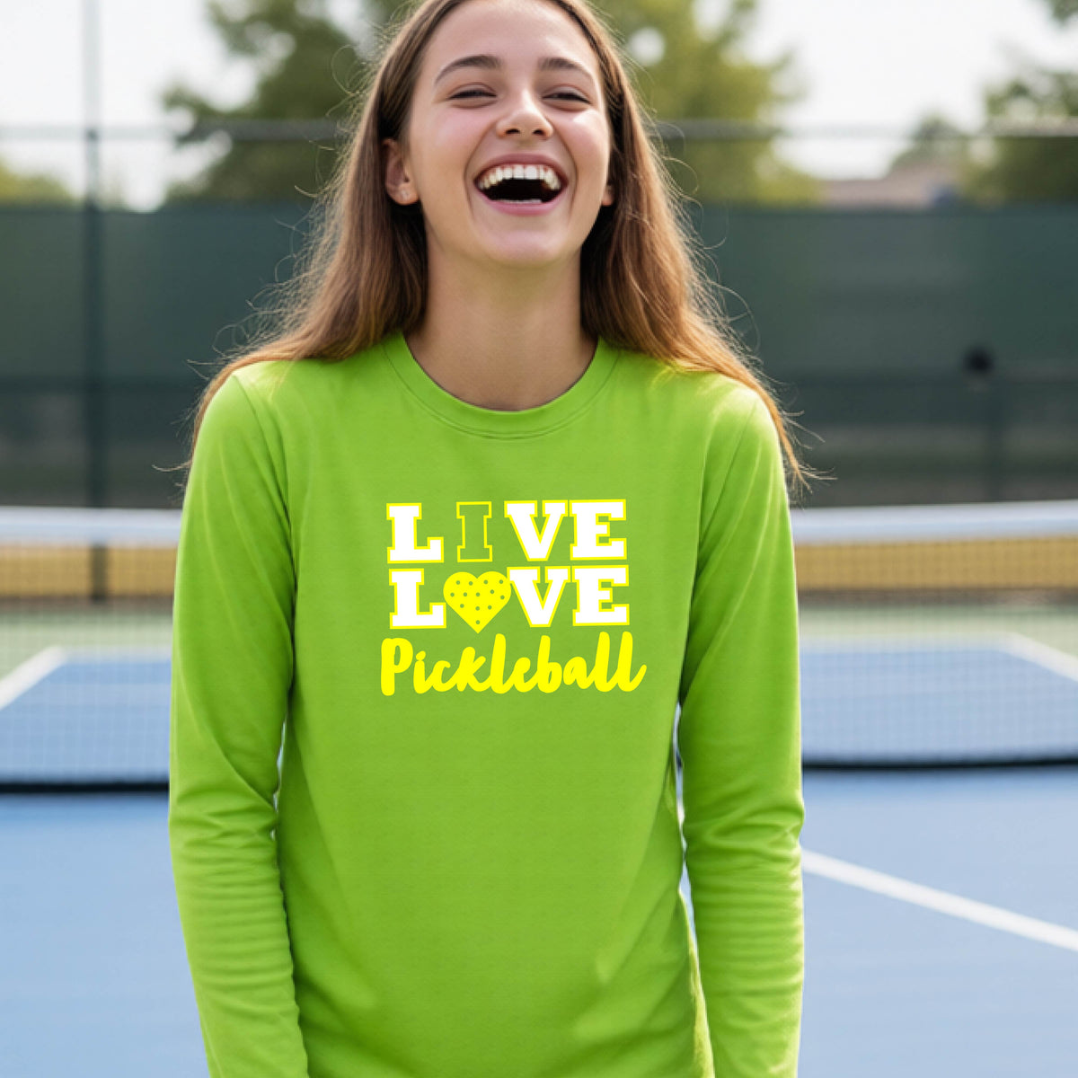 A teenager on a pickleball court wearing a lime green long-sleeve V-neck shirt with a yellow graphic that reads 'LIVE LOVE Pickleball' and a heart-shaped pickleball dot pattern inside the word 'LOVE'.