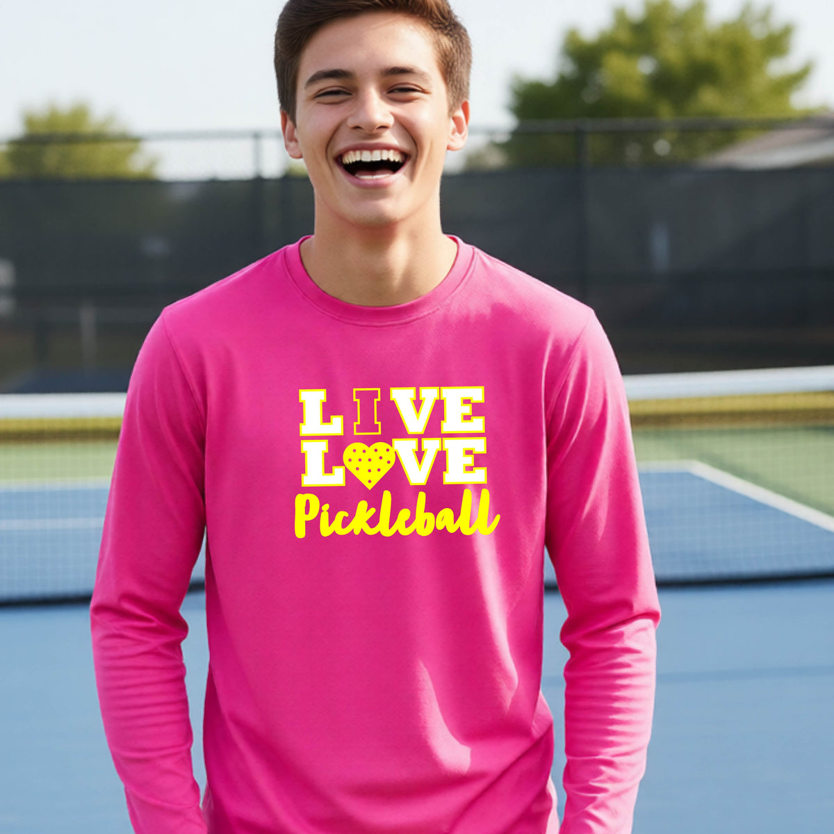 A teenager on a pickleball court wearing a pink long-sleeve V-neck shirt with a yellow graphic that reads 'LIVE LOVE Pickleball' and a heart-shaped pickleball dot pattern inside the word 'LOVE'.