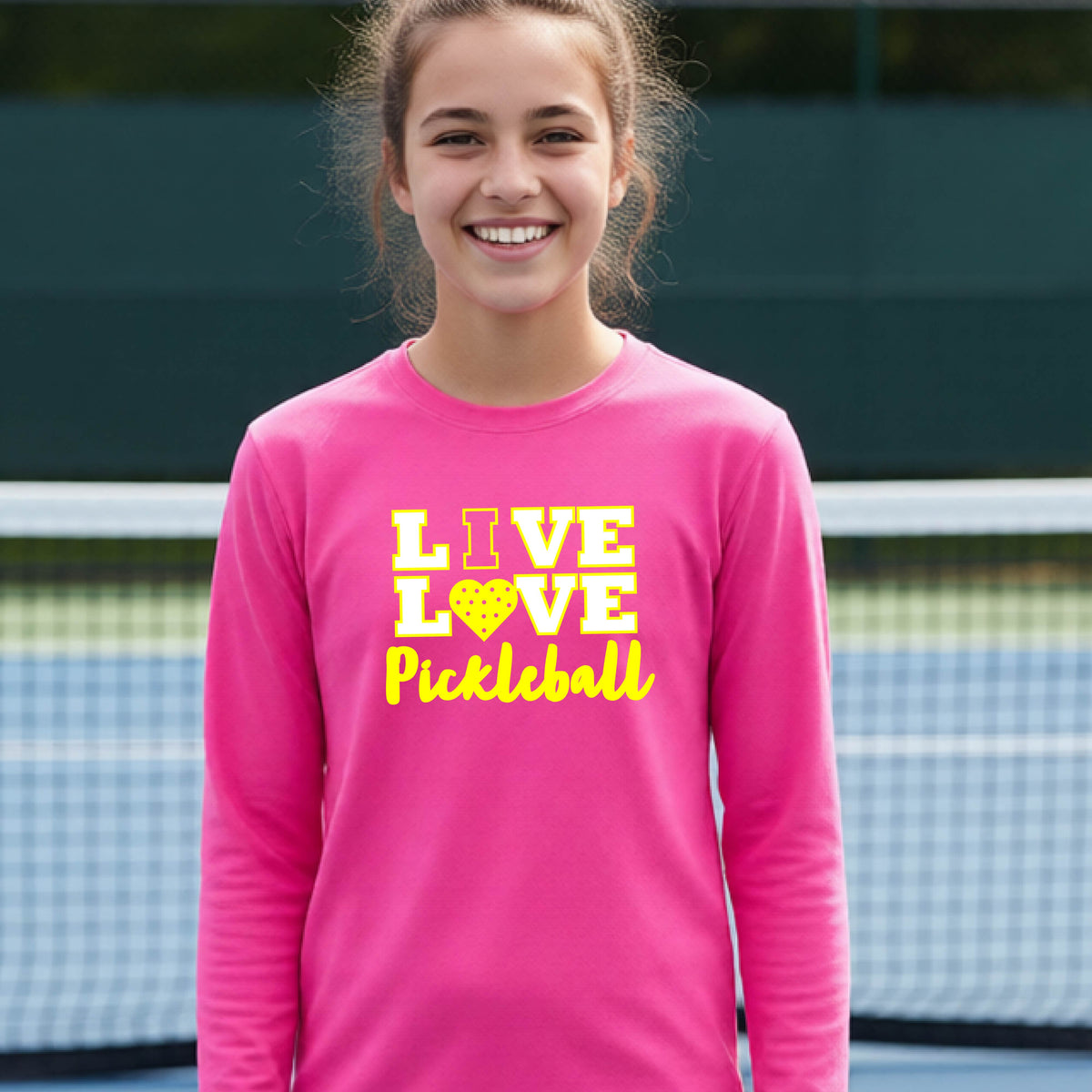 A teenager on a pickleball court wearing a pink long-sleeve V-neck shirt with a yellow graphic that reads 'LIVE LOVE Pickleball' and a heart-shaped pickleball dot pattern inside the word 'LOVE'.