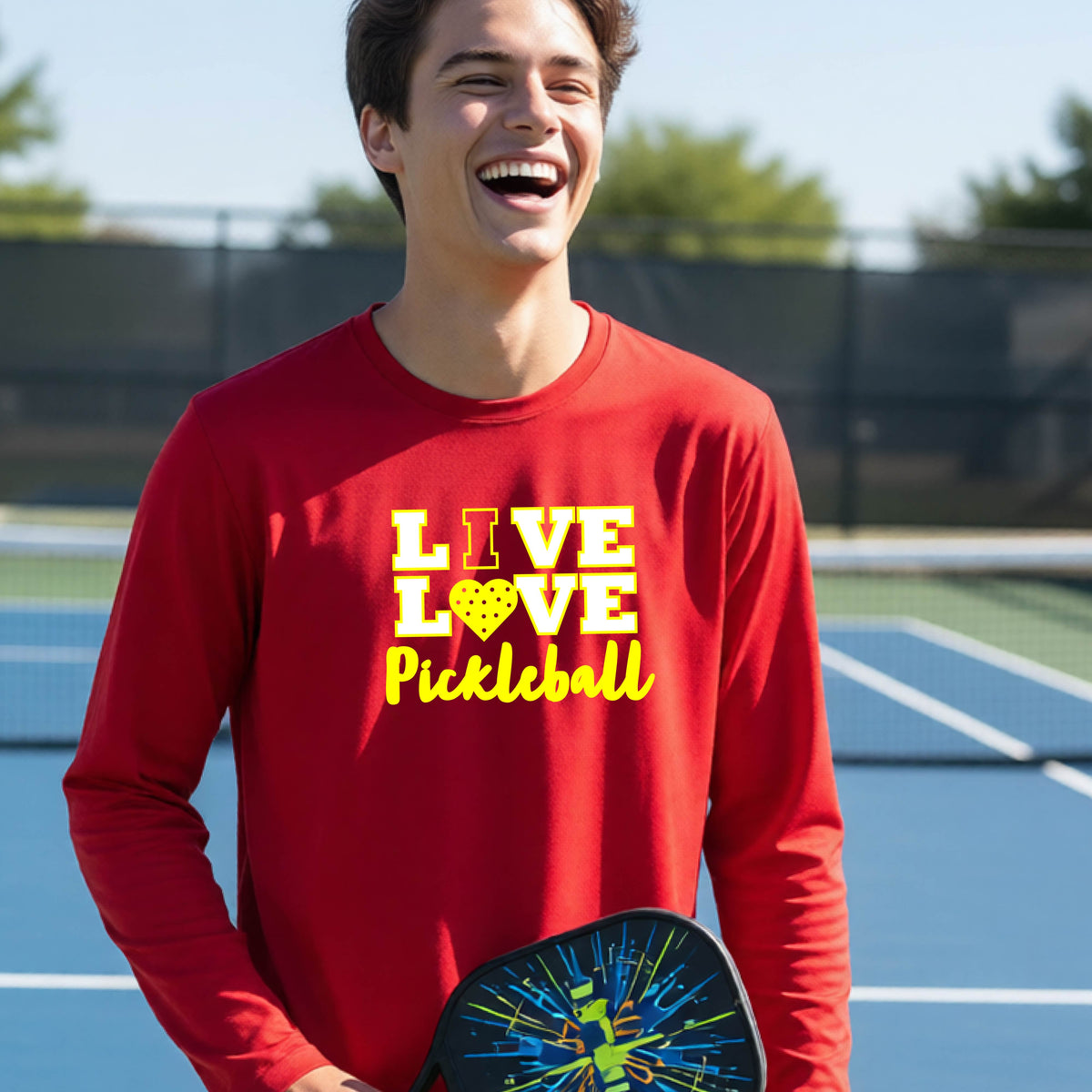 A teenager on a pickleball court wearing a red long-sleeve V-neck shirt with a yellow graphic that reads 'LIVE LOVE Pickleball' and a heart-shaped pickleball dot pattern inside the word 'LOVE'.