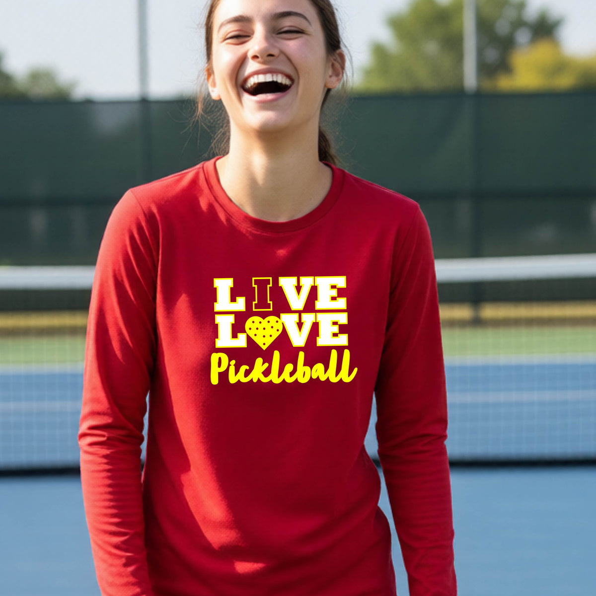 A teenager on a pickleball court wearing a red long-sleeve V-neck shirt with a yellow graphic that reads 'LIVE LOVE Pickleball' and a heart-shaped pickleball dot pattern inside the word 'LOVE'.