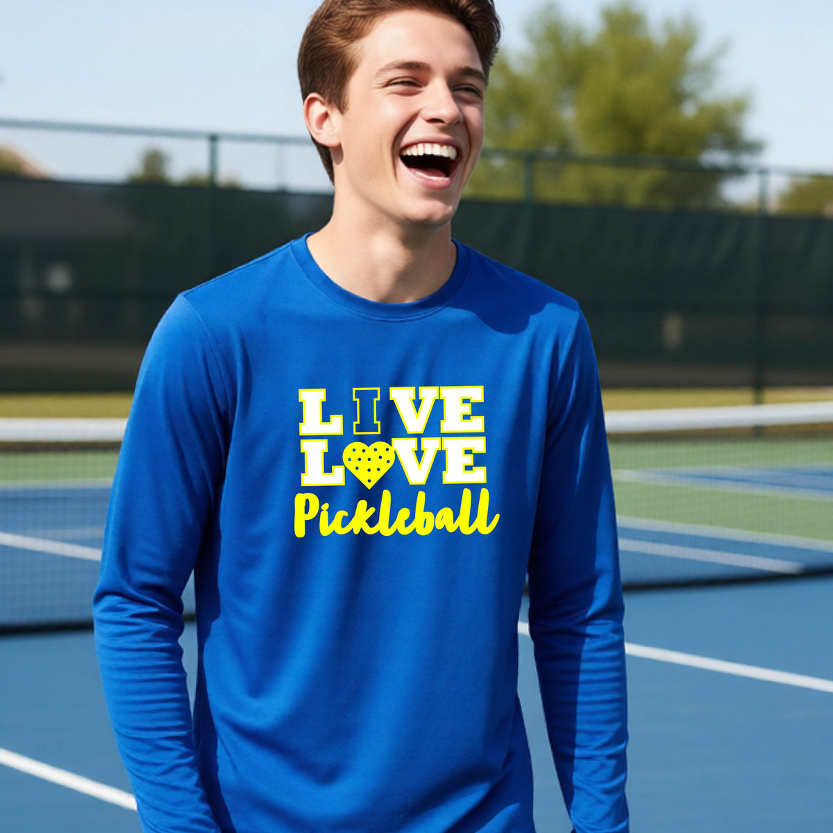 A teenager on a pickleball court wearing a royal blue long-sleeve V-neck shirt with a yellow graphic that reads 'LIVE LOVE Pickleball' and a heart-shaped pickleball dot pattern inside the word 'LOVE'.