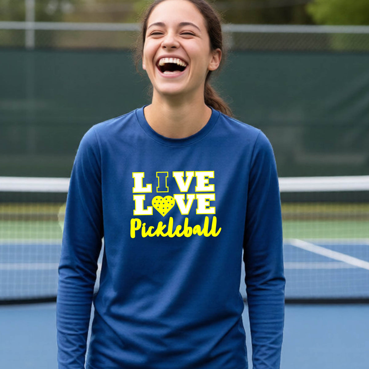 A teenager on a pickleball court wearing a royal blue long-sleeve V-neck shirt with a yellow graphic that reads 'LIVE LOVE Pickleball' and a heart-shaped pickleball dot pattern inside the word 'LOVE'.