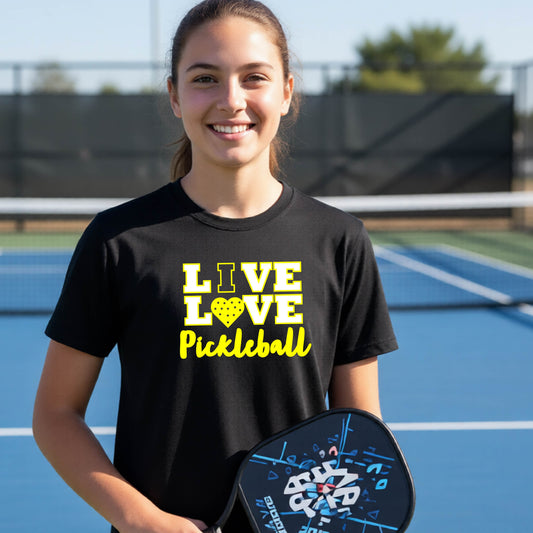 A teenager on a pickleball court wearing a black short-sleeve V-neck shirt with a yellow graphic that reads 'LIVE LOVE Pickleball' and a heart-shaped pickleball dot pattern inside the word 'LOVE'.