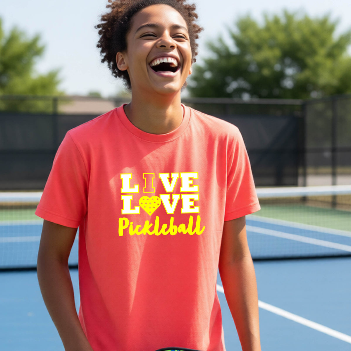 A teenager on a pickleball court wearing a croal orange short-sleeve V-neck shirt with a yellow graphic that reads 'LIVE LOVE Pickleball' and a heart-shaped pickleball dot pattern inside the word 'LOVE'.