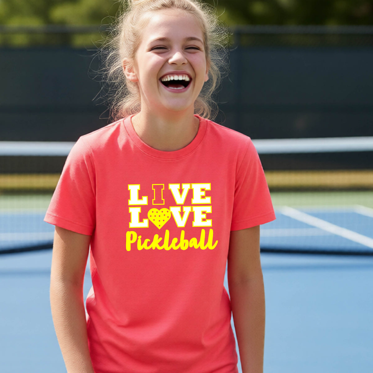 A teenager on a pickleball court wearing a coral orange short-sleeve V-neck shirt with a yellow graphic that reads 'LIVE LOVE Pickleball' and a heart-shaped pickleball dot pattern inside the word 'LOVE'.