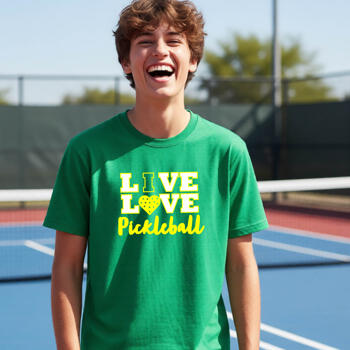 A teenager on a pickleball court wearing a green short-sleeve V-neck shirt with a yellow graphic that reads 'LIVE LOVE Pickleball' and a heart-shaped pickleball dot pattern inside the word 'LOVE'.