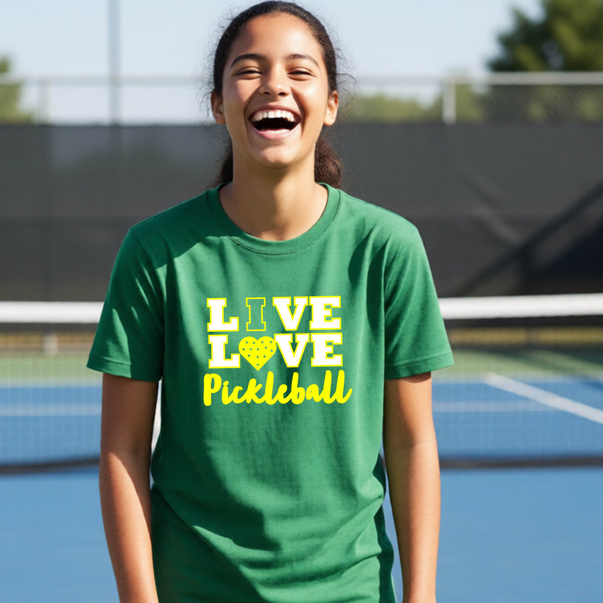 A teenager on a pickleball court wearing a green short-sleeve V-neck shirt with a yellow graphic that reads 'LIVE LOVE Pickleball' and a heart-shaped pickleball dot pattern inside the word 'LOVE'.
