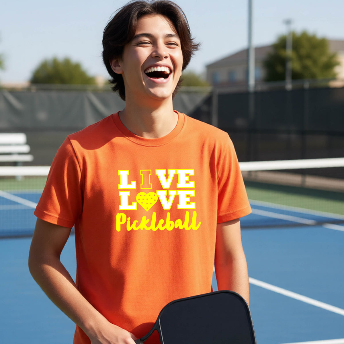 A teenager on a pickleball court wearing a orange short-sleeve V-neck shirt with a yellow graphic that reads 'LIVE LOVE Pickleball' and a heart-shaped pickleball dot pattern inside the word 'LOVE'.