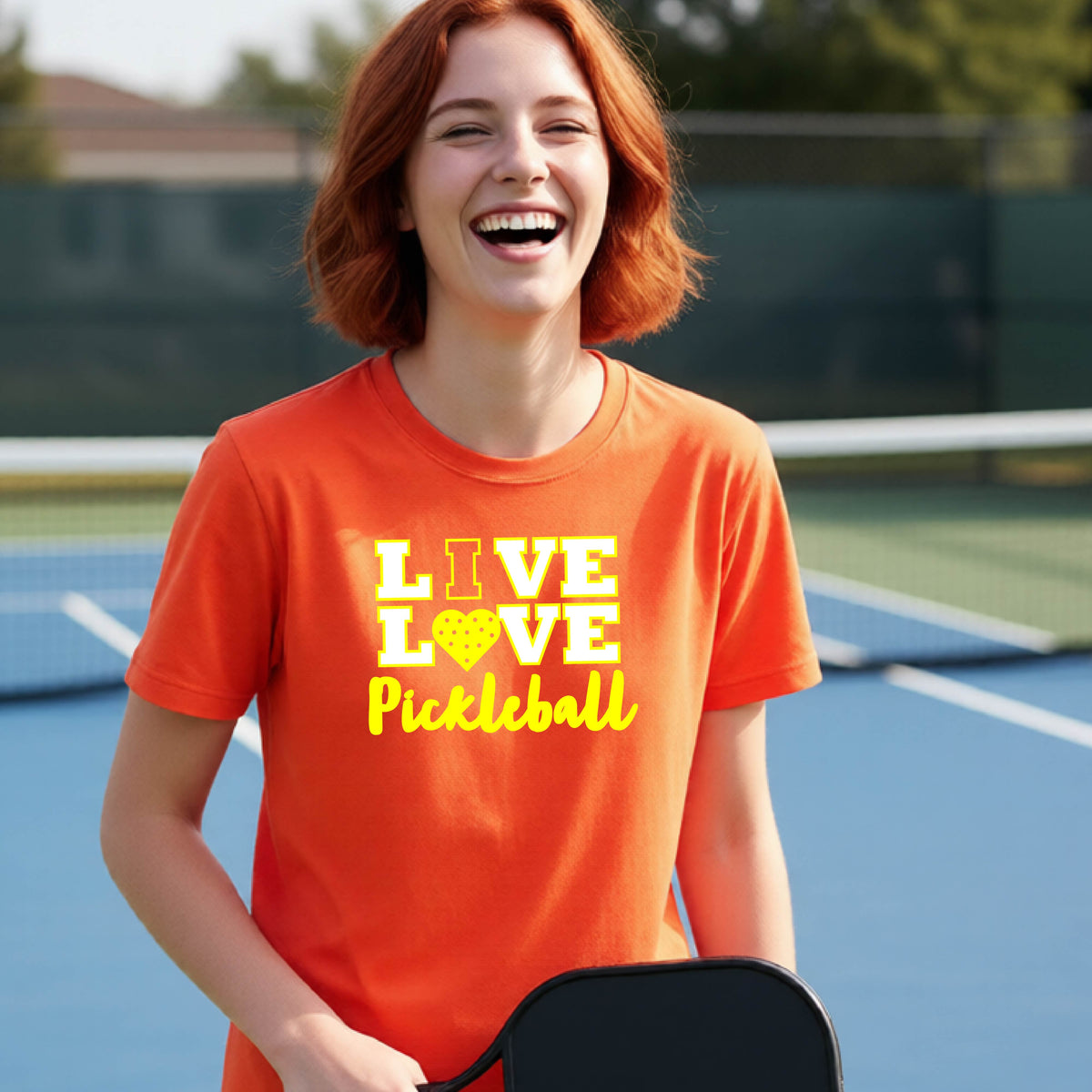 A teenager on a pickleball court wearing a orange short-sleeve V-neck shirt with a yellow graphic that reads 'LIVE LOVE Pickleball' and a heart-shaped pickleball dot pattern inside the word 'LOVE'.