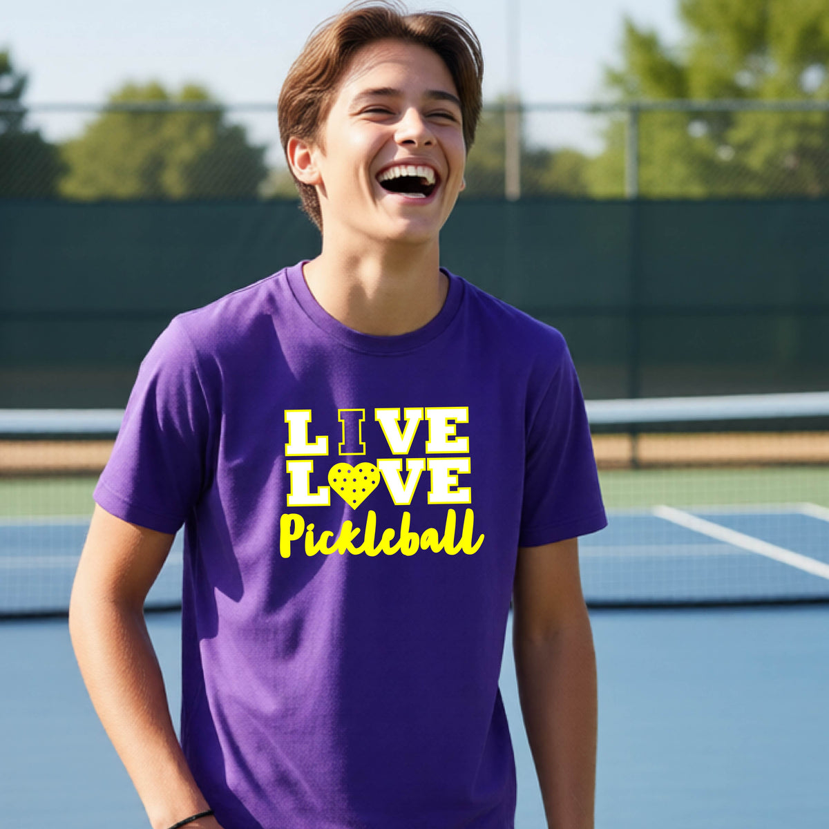 A teenager on a pickleball court wearing a purple short-sleeve V-neck shirt with a yellow graphic that reads 'LIVE LOVE Pickleball' and a heart-shaped pickleball dot pattern inside the word 'LOVE'.