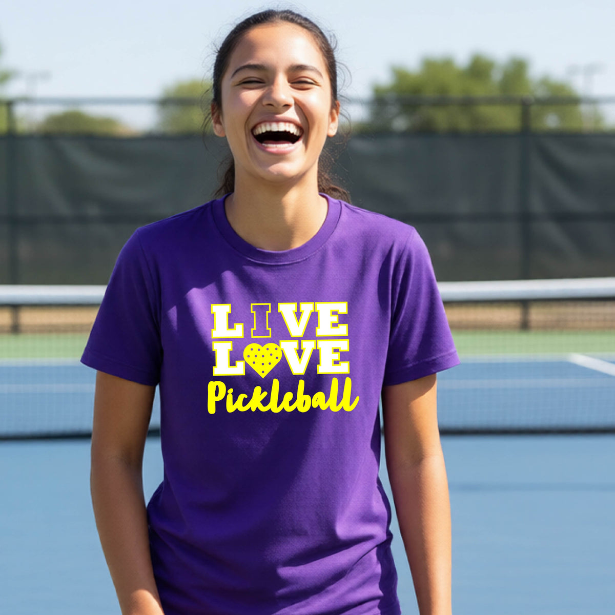 A teenager on a pickleball court wearing a purple short-sleeve V-neck shirt with a yellow graphic that reads 'LIVE LOVE Pickleball' and a heart-shaped pickleball dot pattern inside the word 'LOVE'.