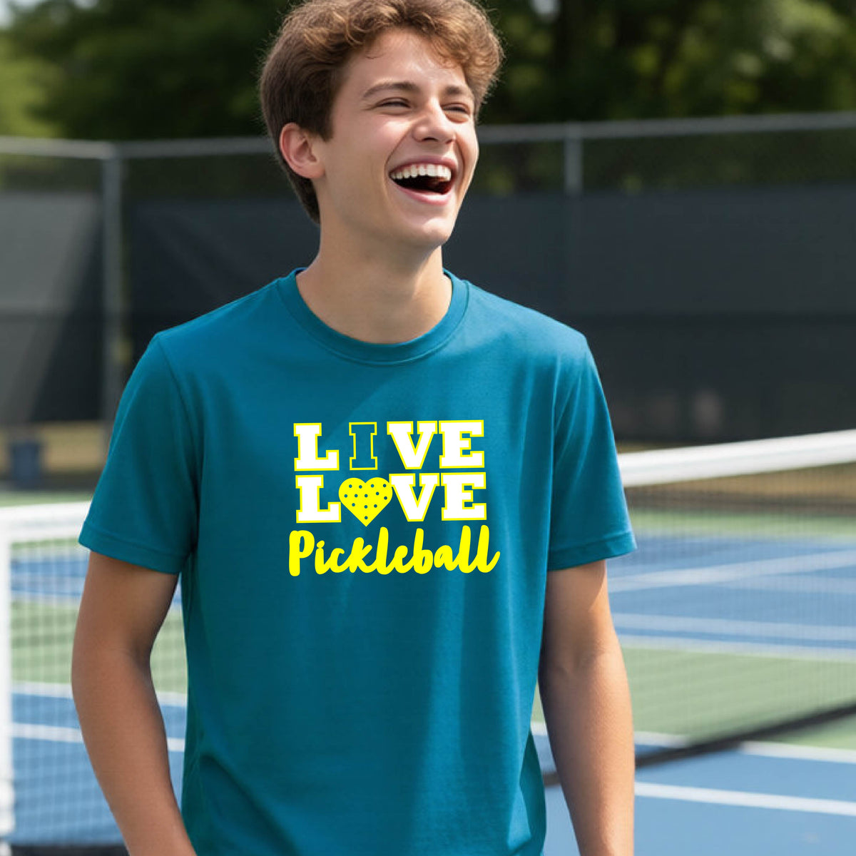 A teenager on a pickleball court wearing a teal blue short-sleeve V-neck shirt with a yellow graphic that reads 'LIVE LOVE Pickleball' and a heart-shaped pickleball dot pattern inside the word 'LOVE'.