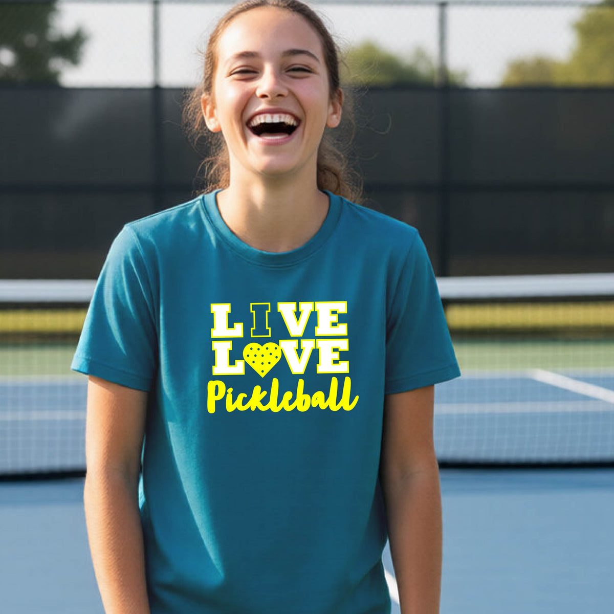 A teenager on a pickleball court wearing a teal blue short-sleeve V-neck shirt with a yellow graphic that reads 'LIVE LOVE Pickleball' and a heart-shaped pickleball dot pattern inside the word 'LOVE'.